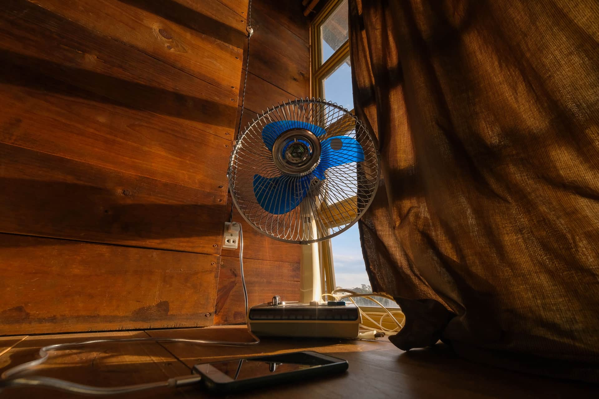 A tower fan running beside an open window in a lived-in bedroom in late afternoon light